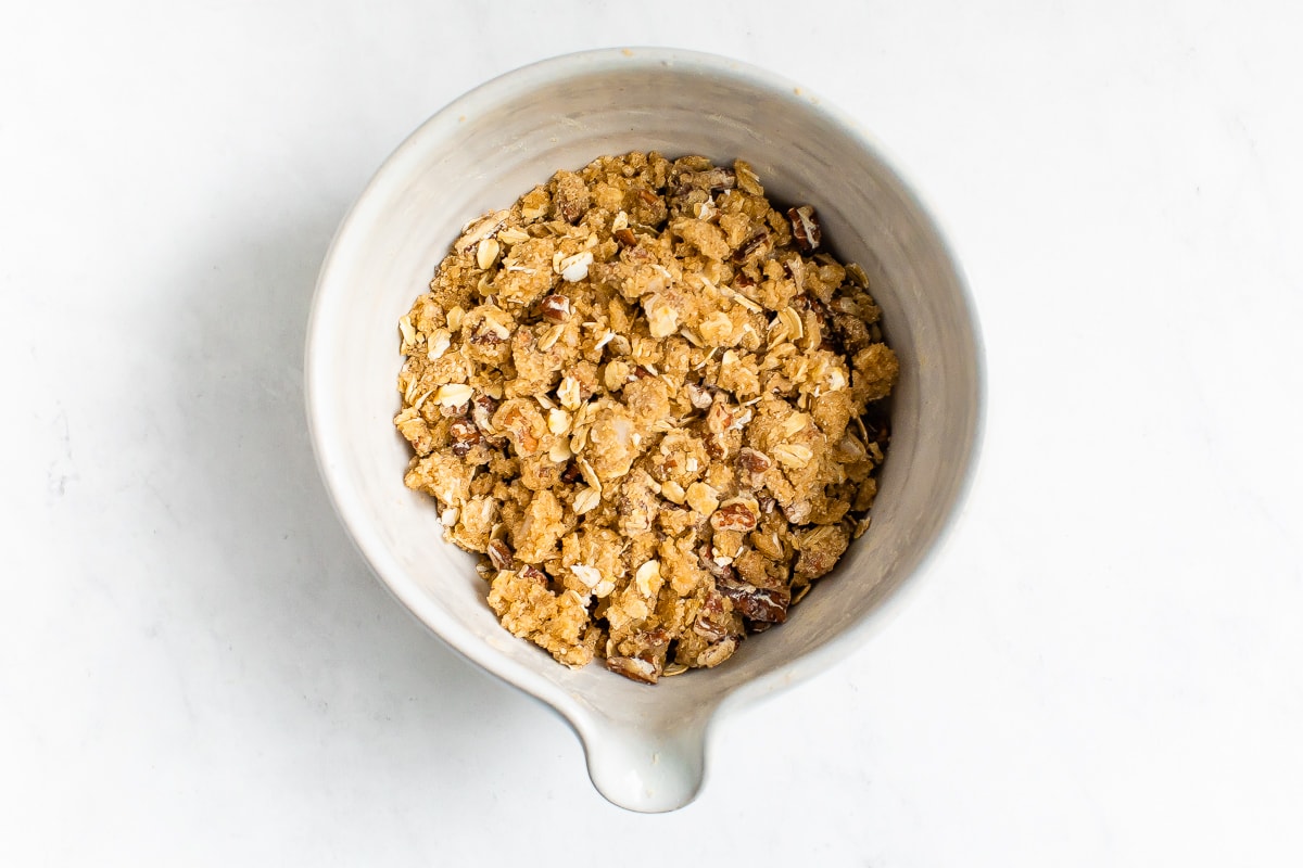 A ceramic mixing bowl containing oats, brown sugar, pecans, oat flour and coconut oil mixed together to make a topping for casserole.