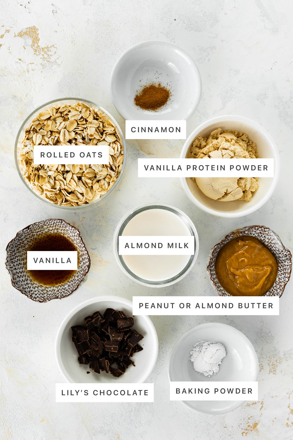 Ingredients measured out to make Air Fryer Baked Oats: rolled oats, cinnamon, vanilla protein powder, vanilla, almond milk, peanut butter, Lily's chocolate and baking powder.
