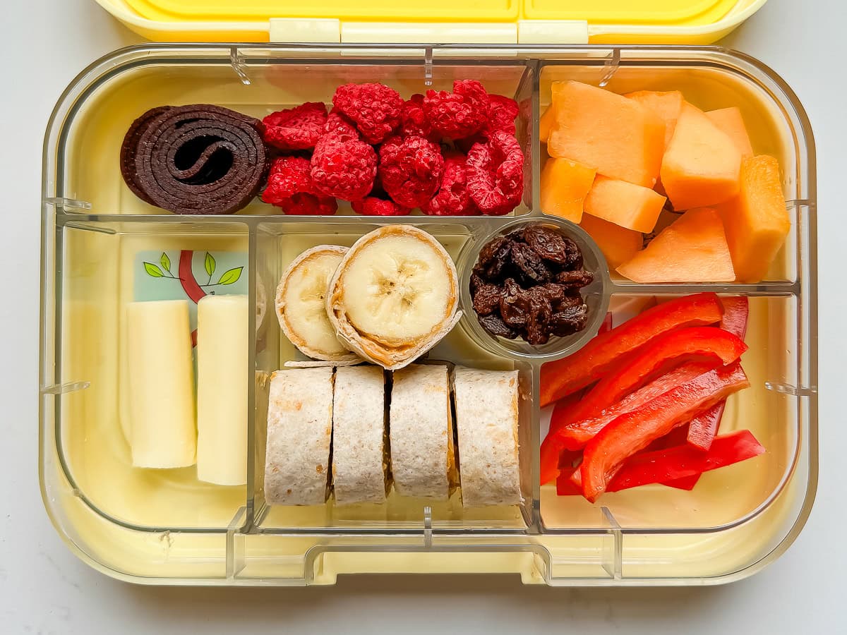 A kids lunch box with banana sushi, a cheese stick, bell pepper slices, melon, freeze dried raspberries and a natural fruit roll.