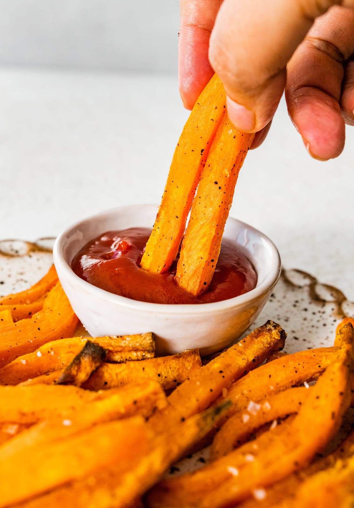 A woman's hand dipping two sweet potato fries in a small white bowl of ketchup.